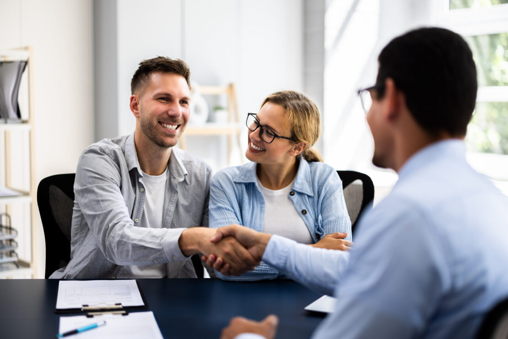 Two men and a woman shaking hands in an office, with papers and pens on the table between them