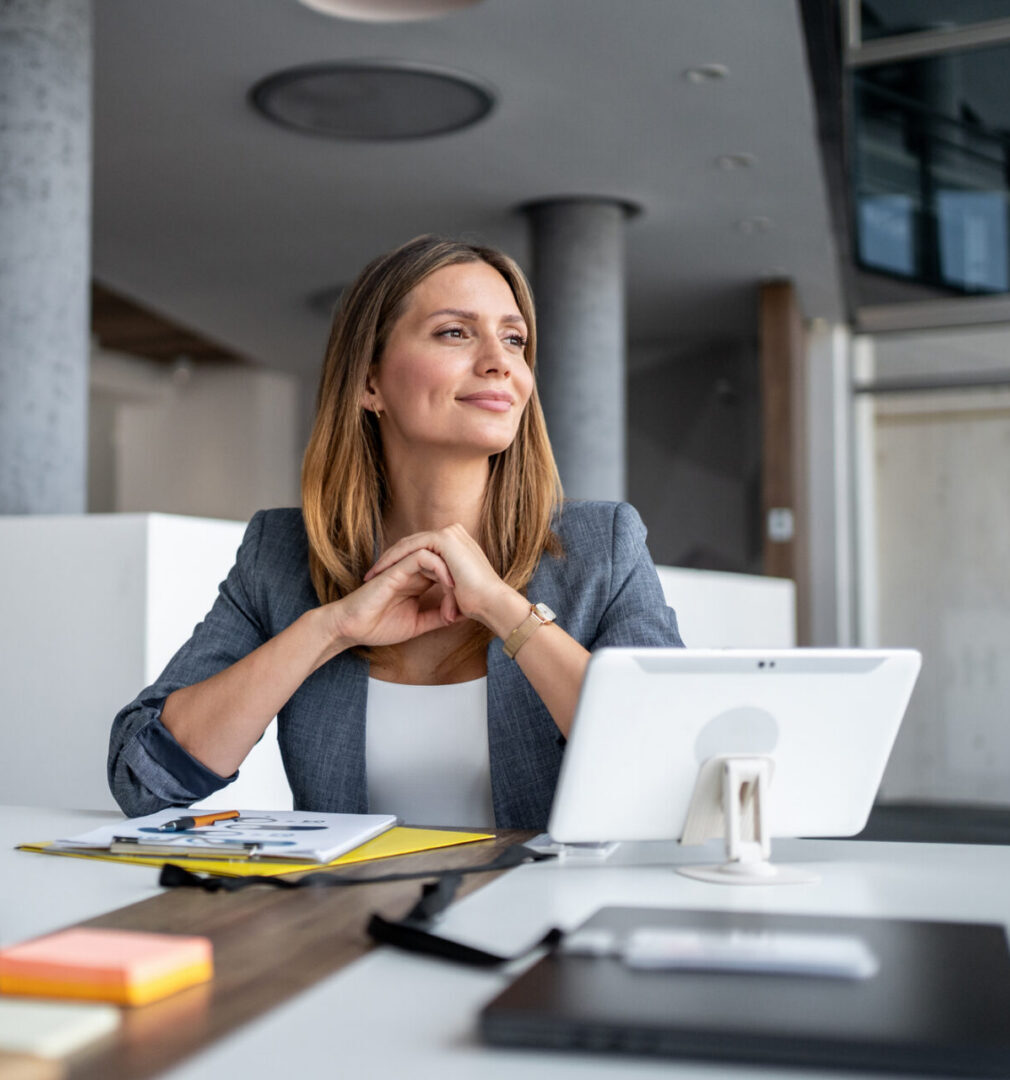 Businesswoman sitting at a modern office desk, smiling and gazing thoughtfully into the distance while contemplating future business plans and aspirations for success