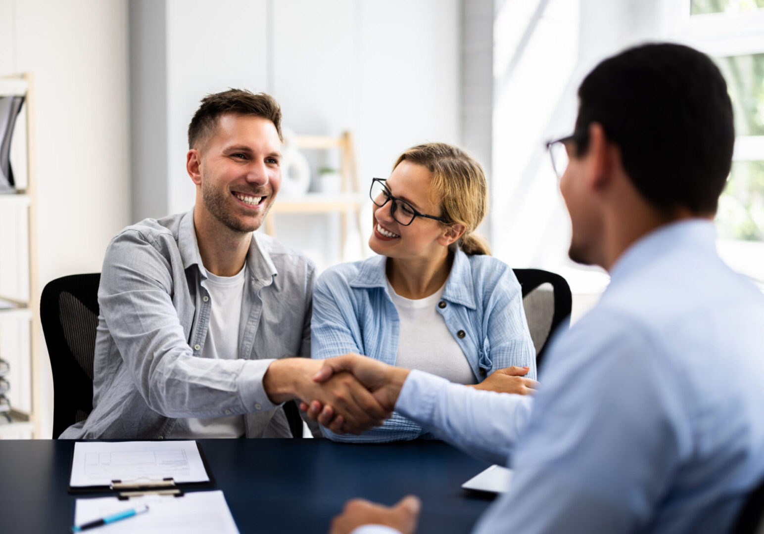 Two men and a woman shaking hands in an office, with papers and pens on the table between them